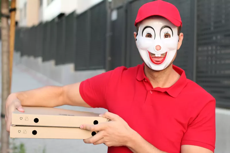 Image: A man wearing a strange, doll-like mask holds a few pizza boxes at his side