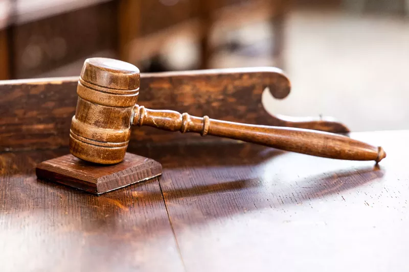 Image: A judge's gavel sits on a wooden desk in a courtroom.