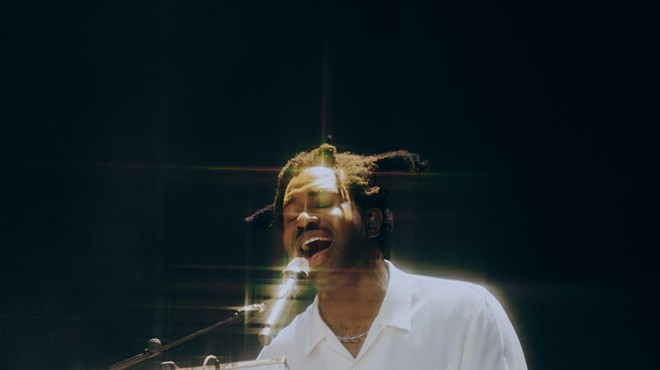 color photo of a white-shirted, dreadlocked Sampha singing into a microphone against a black background