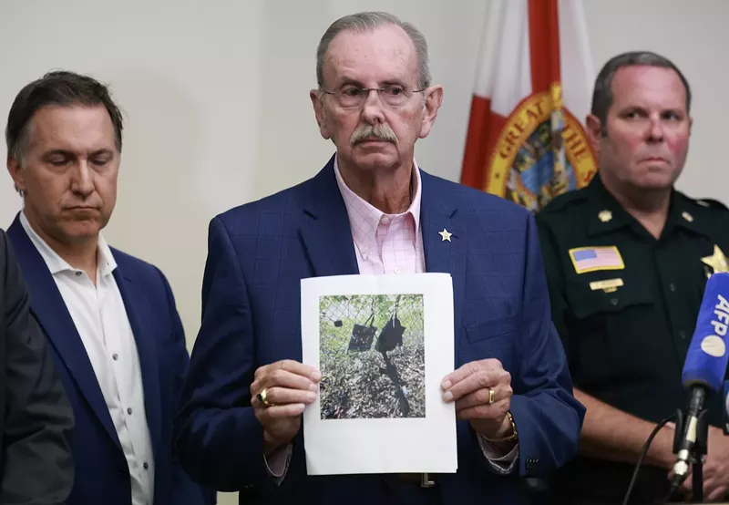 Image: photo of a gray-haired, mustachioed man wearing wire-rimmed glasses looking somber as he holds up a color printout showing a rifle and other items near a chain-link fence