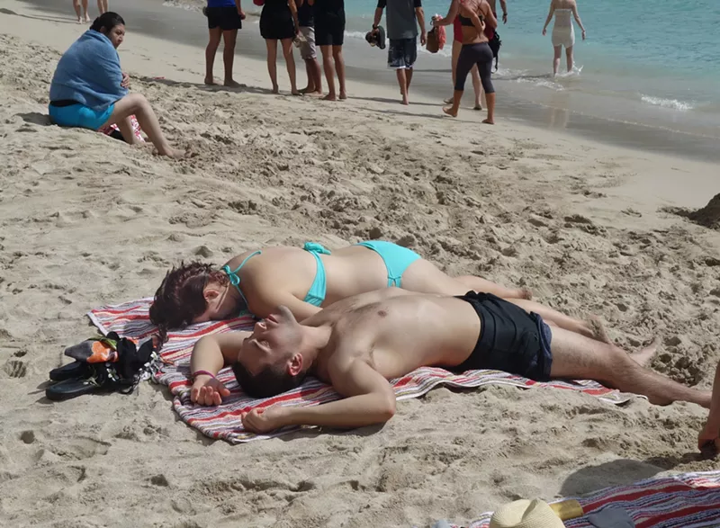Image: A couple relaxing on sandy shores, lying side by side on a colorful striped towel, soaking up the sun as beachgoers stroll nearby
