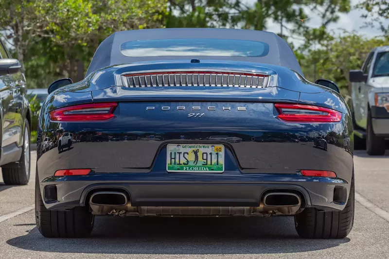 Image: Rear end of a dark blue convertible Porsche 911 in a parking lot with a golfer on the license plate and dual exhausts