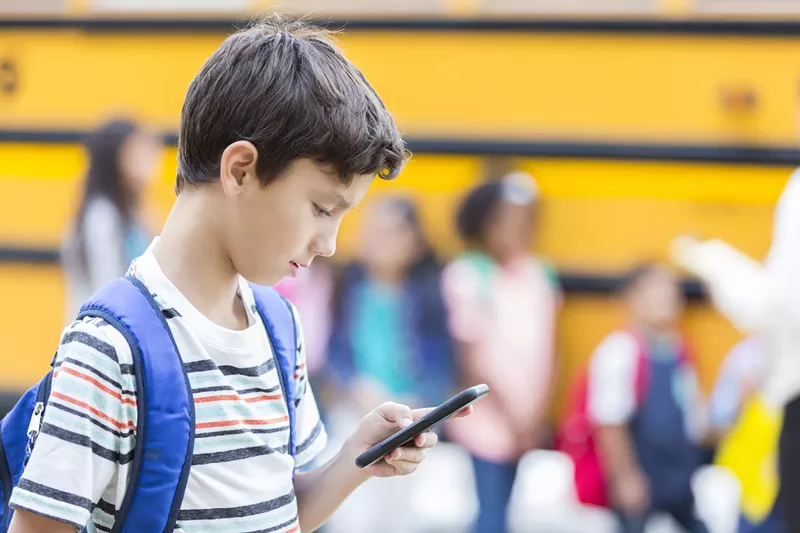 Image: An elementary-school age boy with a blue backpack looks down at his cellphone in front of a school bus