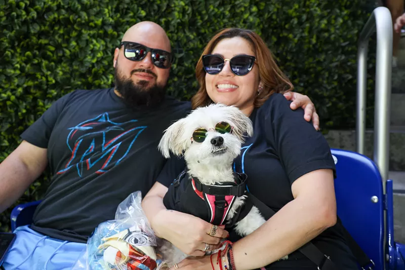 Image: Two people pose with their dog. Both people and the dog are wearing shades and baseball tees