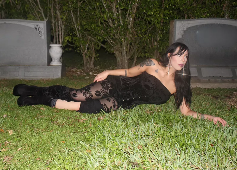 Image: a woman in a black corset and black floral tights lays in the grass at a cemetry