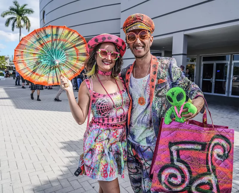 Image: a woman and man in painted colorful clothing poses in front of a convention center. the woman holds a painted parasol