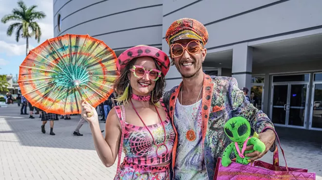 a woman and man in painted colorful clothing poses in front of a convention center. the woman holds a painted parasol
