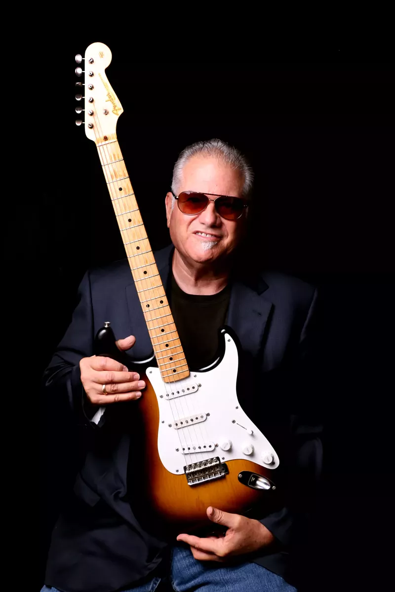 Image: A man with slicked-back hair wearing sunglasses and holding an electric guitar
