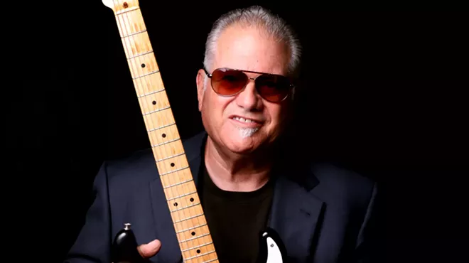 A man with slicked-back hair wearing sunglasses and holding an electric guitar