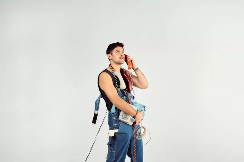 Image: Portrait of a young man against a white backdrop holding many old telephones.