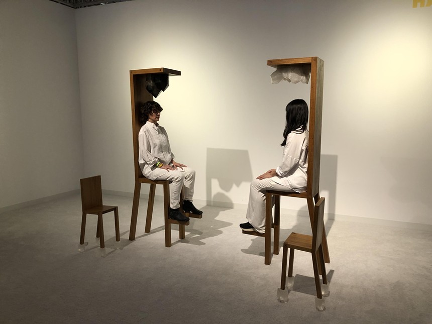 Two women face each other sitting in wooden chairs by artist Marina Abramovic