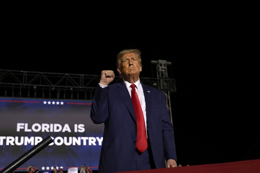 Donald Trump holds up his fist on stage, in front of a "Florida Is Trump Country" screen