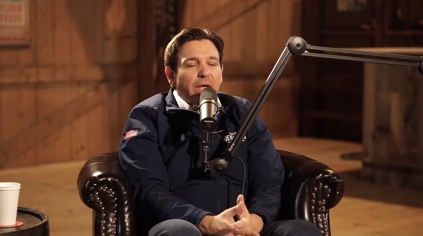 Florida Gov. Ron DeSantis hunching in front of a microphone in a leather upholstered chair looking like a spoiled little boy