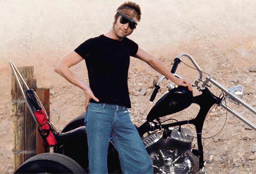 color photo of the ex-Beatle, wearing aviator sunglasses and a headband, a black T-shirt, and blue jeans, leaning against a three-wheeled black chopper on a dusty landscape