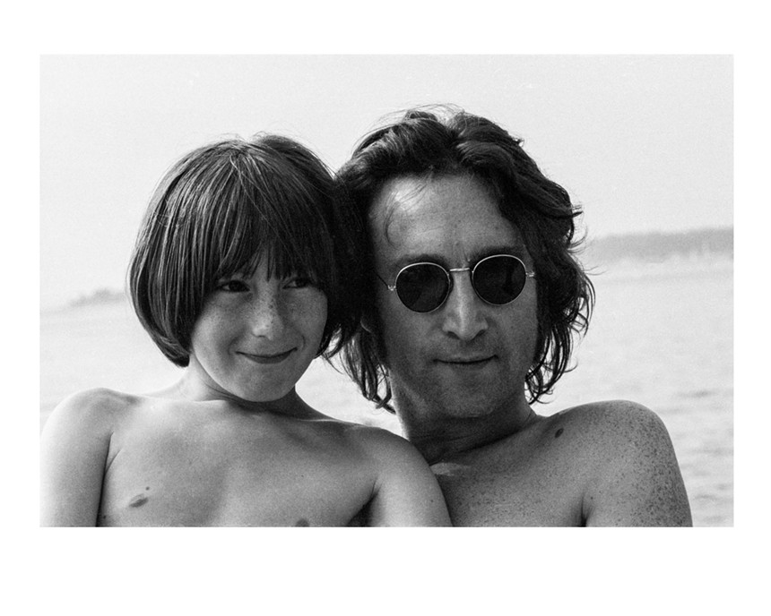 Black-and-white photo of Julian and John Lennon, shirtless. Julian is smiling, Lennon is wearing his trademark round-lensed sunglasses.