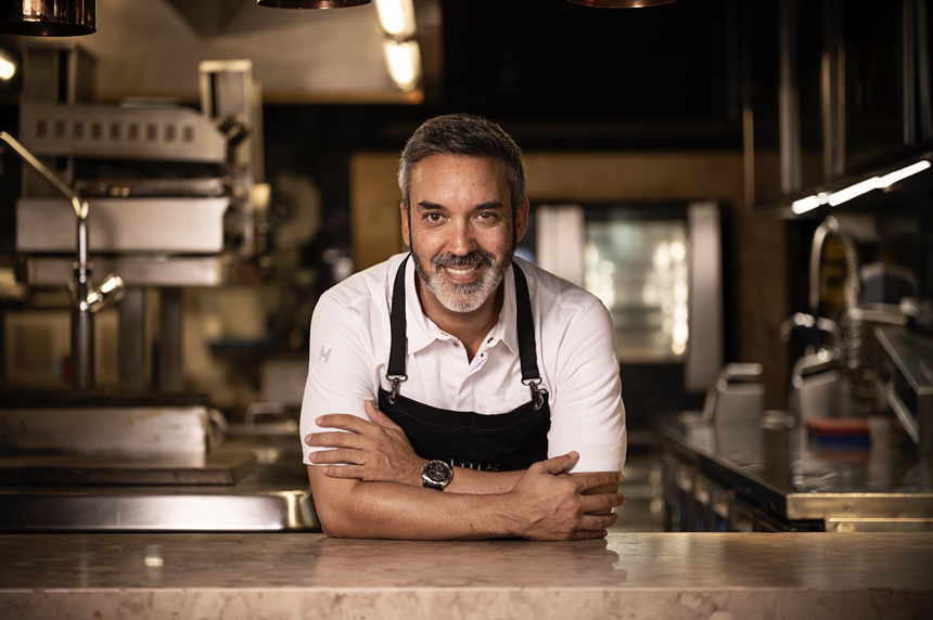 A chef poses with a black apron against a marble table