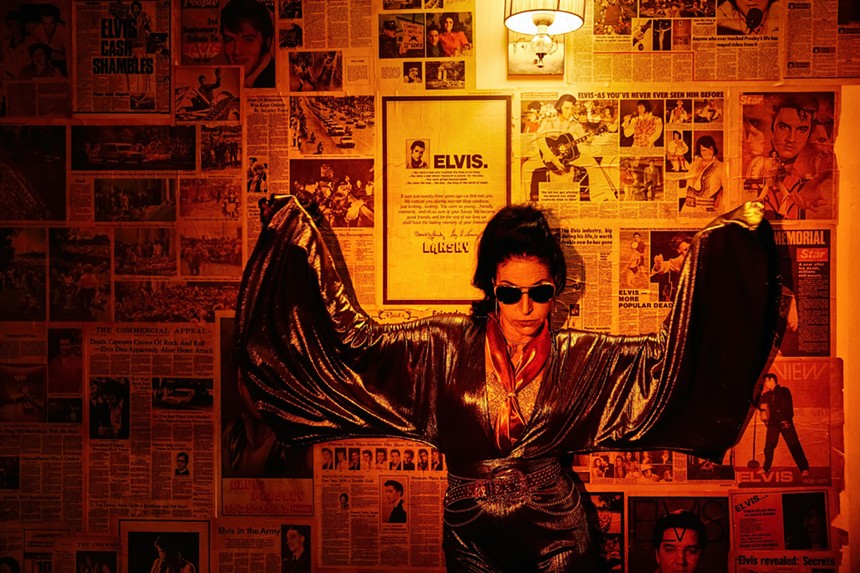 Susie K. Taylor in a gold jumpsuit and sunglasses against a wall plastered with Elvis newspaper clippings