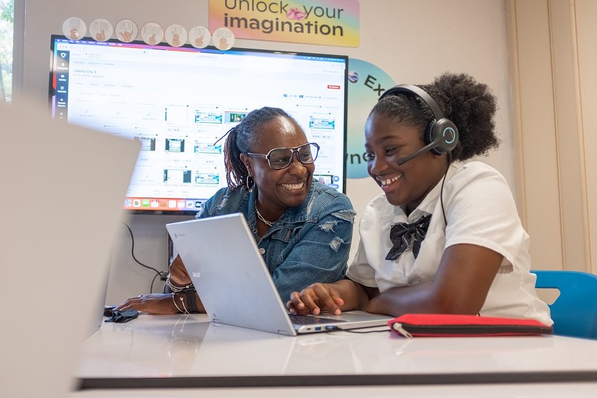 A teacher smiles next to a student, who is wearing headphones while working on a laptop