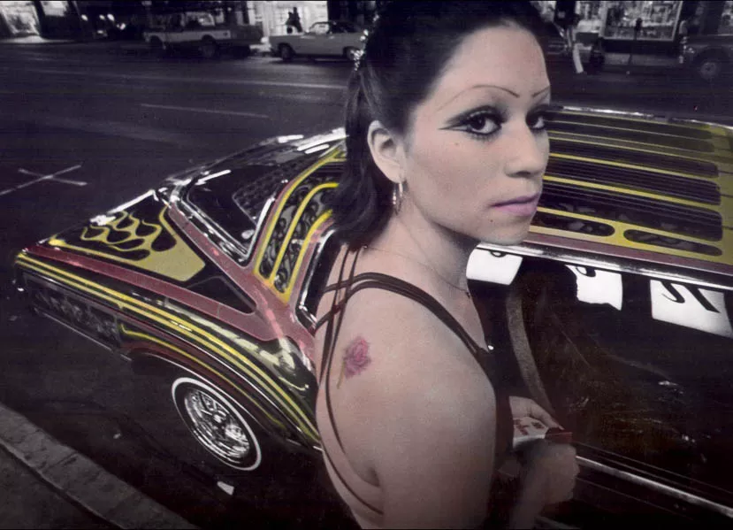 A Chicana woman poses in front of a car.