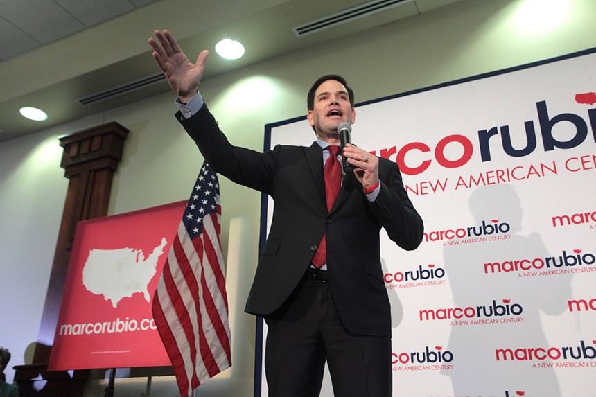 Sen. Marco Rubio speaks on a conference stage with his right hand extended upwards