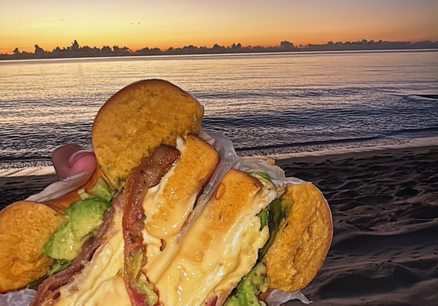 a bacon egg and cheese sandwich in front of a sunrise at the beach