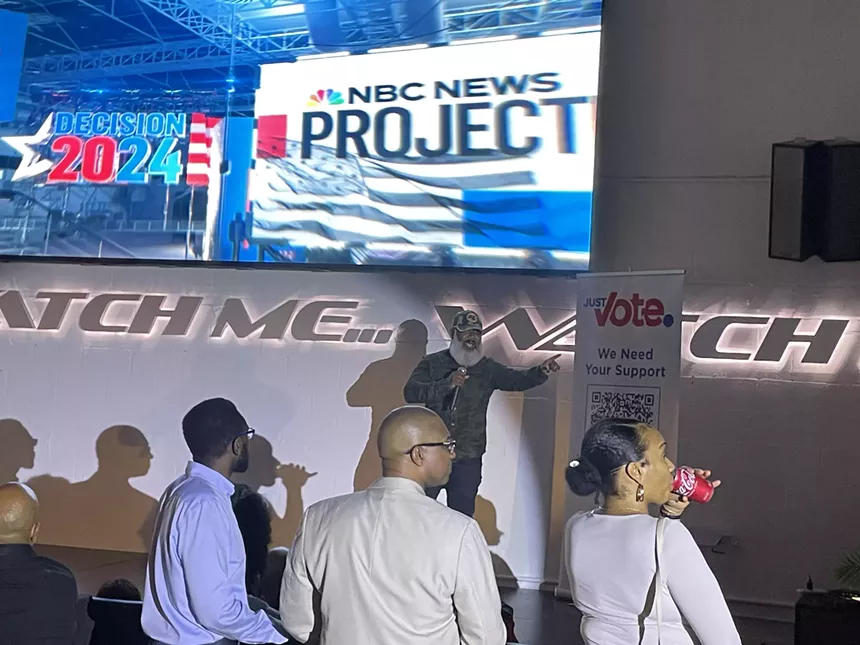 Shareef Nash speaking to attendees at Just Vote Period watch party with "Decision 2024" displayed on screen behind him