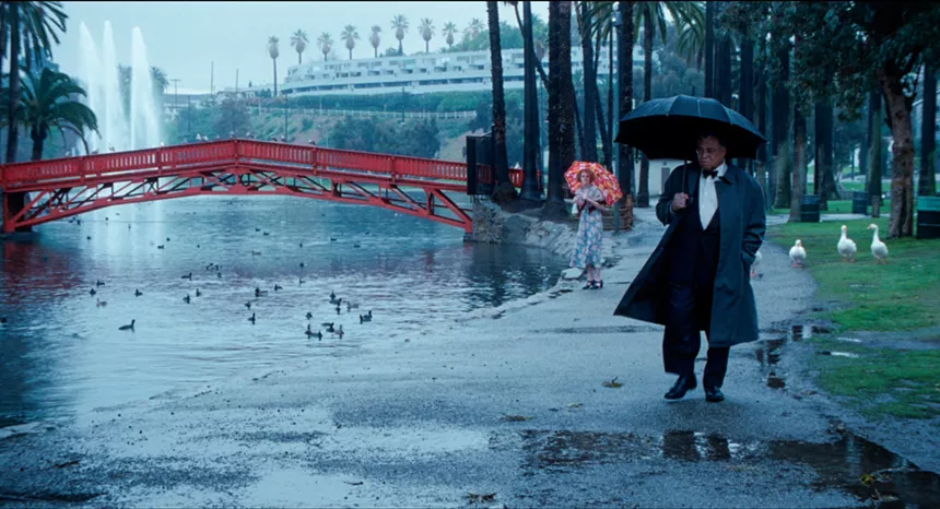 A man holding an umbrella walks next to a pond and a red bridge on a gray rainy day. A woman stands behind him holding a red patterned umbrella. Ducks swim in the pond on his left. Geese stand behind him on the right.