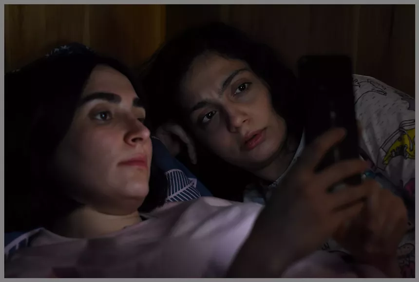 Two young women sit in the dark looking at a phone. The glare lights up their faces