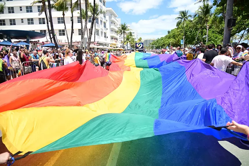 a huge Pride flag is paraded down a parade route with spectators on each side