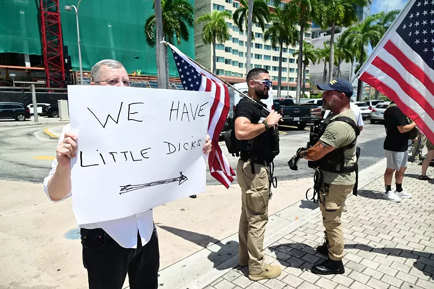 An elderly man holds up a Sharpied white sign reading "we have little dicks" with an arrow pointing right. Two law enforcement officers stand to the right of the sign.