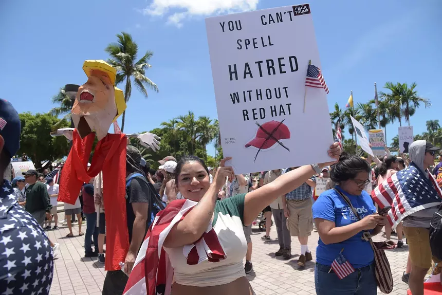A protester holds up a sign reading, "You can't spell hatred without red hat." A picture of a red hat is crossed out below the text