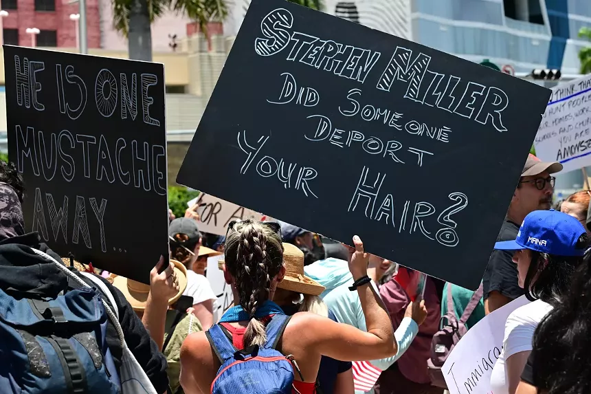 Protesters hold signs reading, "He is one mustache away," and "Stephen Miller, did someone deport your hair?"