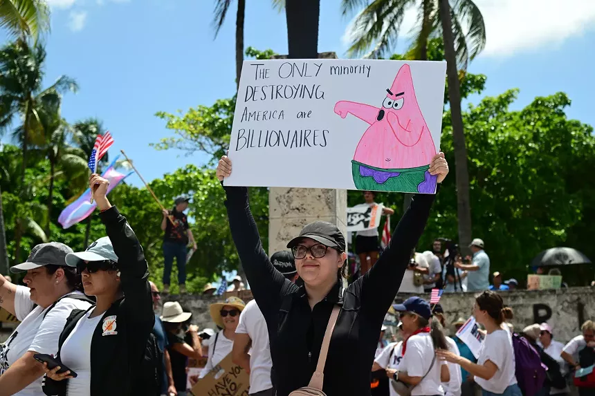 A protester holds up a sign depicting Patrick Star booing next to text reading, "The ONLY minority DESTROYING America are BILLIONAIRES"