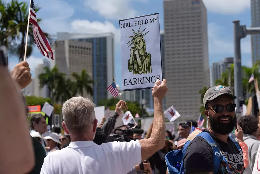 A man holds up a sign depicting the Statue of Liberty removing her earrings. The text on the sign reads, "Girl, hold my earrings."