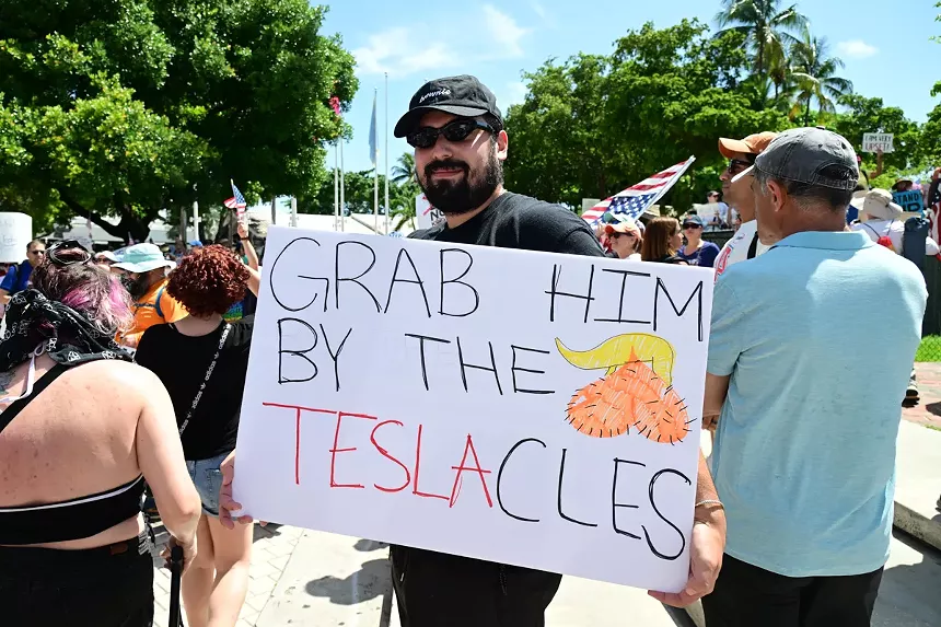 Protester holds up a sign reading "Grab him by the Teslacles" with a drawing of an orange scrotum with Donal Trump's hair