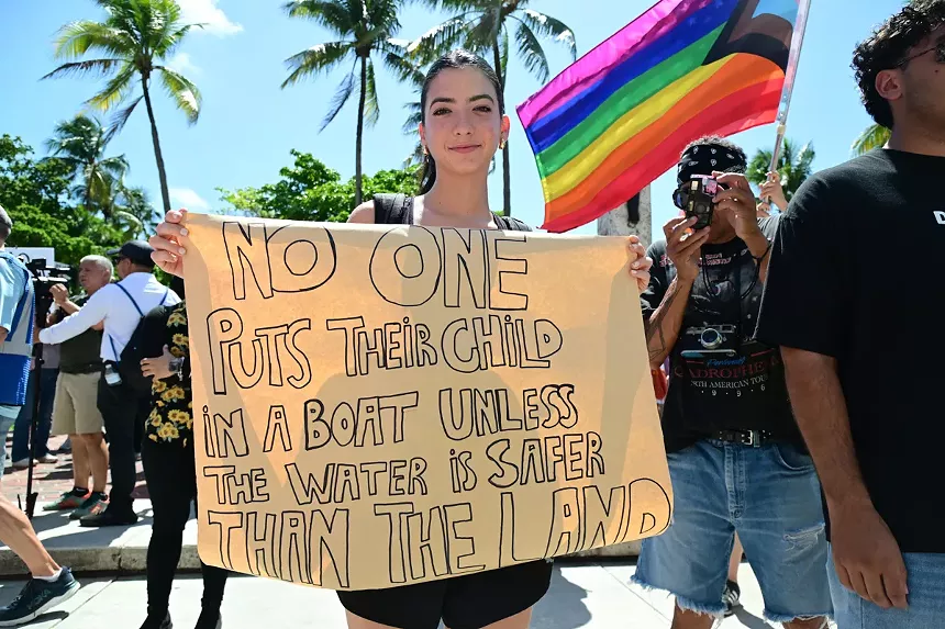 Protester holds up a sign reading, "No one puts their child on a boat unless the water is safer than the land."