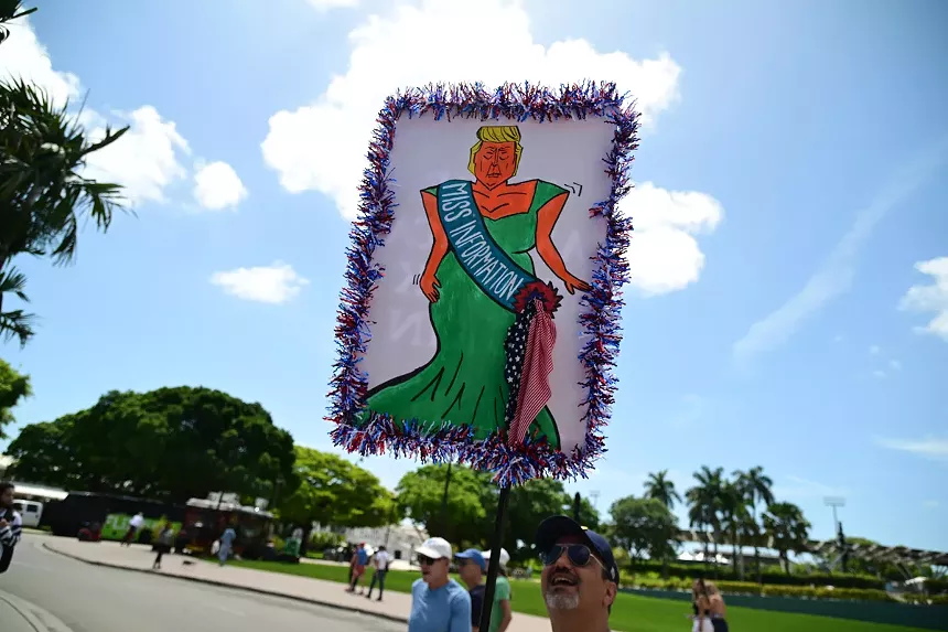 A protest sign depicts Donald Trump as a pageant queen with a sash reading "Miss Information"