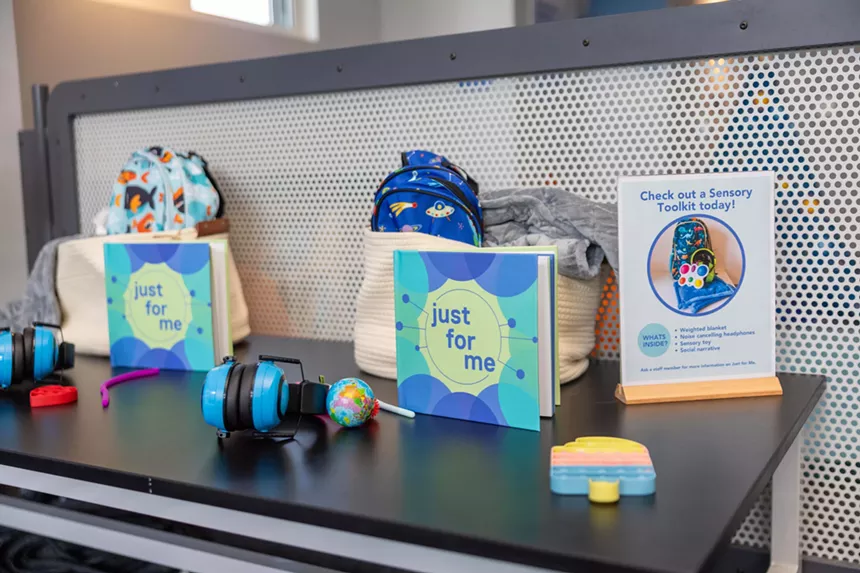 A table with backpacks and children's books that read, "Just for Me." A sign on the table reads, "Check out a Sensory Toolkit today!"