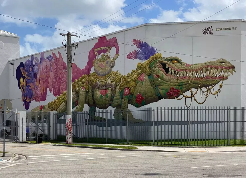a white building displays a mural with an alligator wearing regal decor, a tea kettle, and feathers