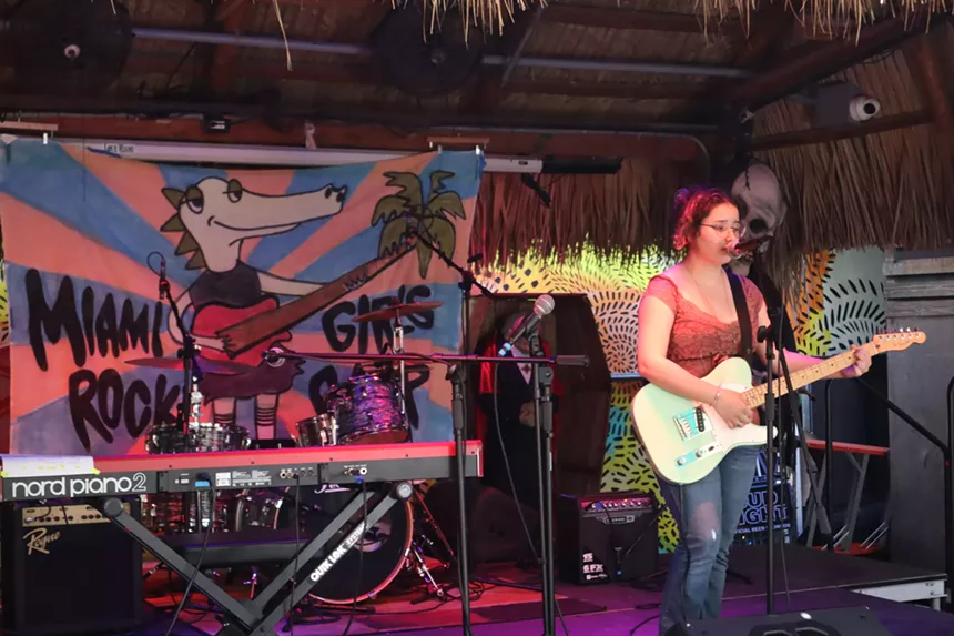 A young girl plays guitar on stage. A sign behind her reads, "Miami Girls Rock Camp"