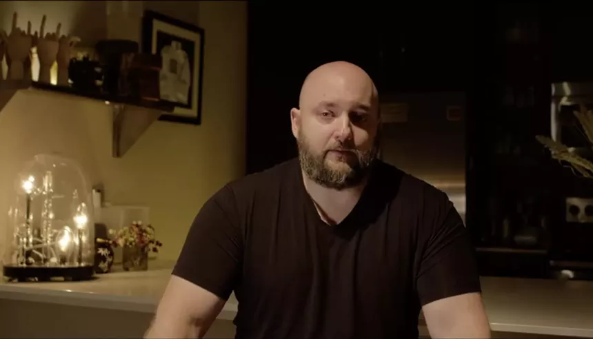 screenshot of a man with a shaved head and a full beard, seated at a kitchen counter