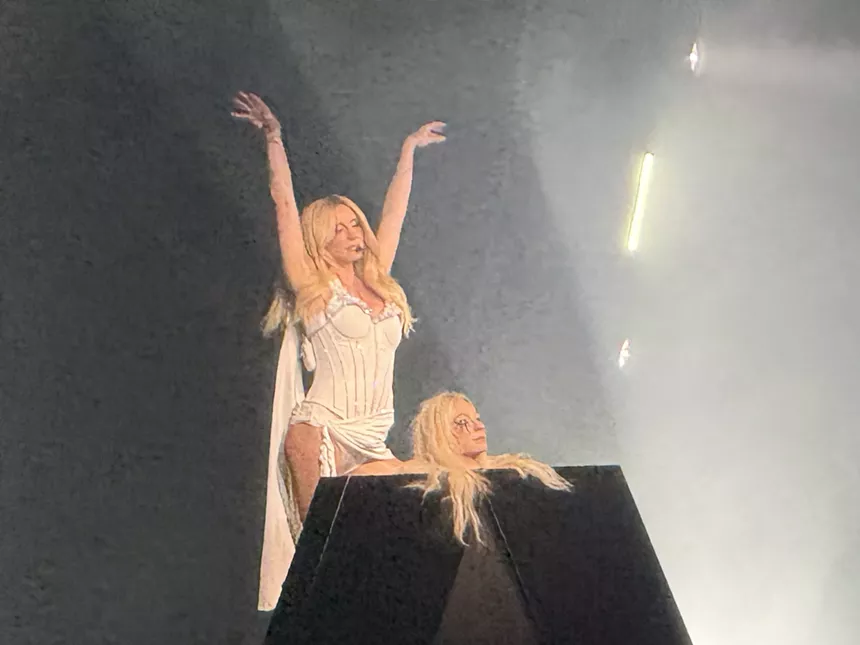 Photo of Kesha on stage wearing white and wielding an effigy of her own severed head