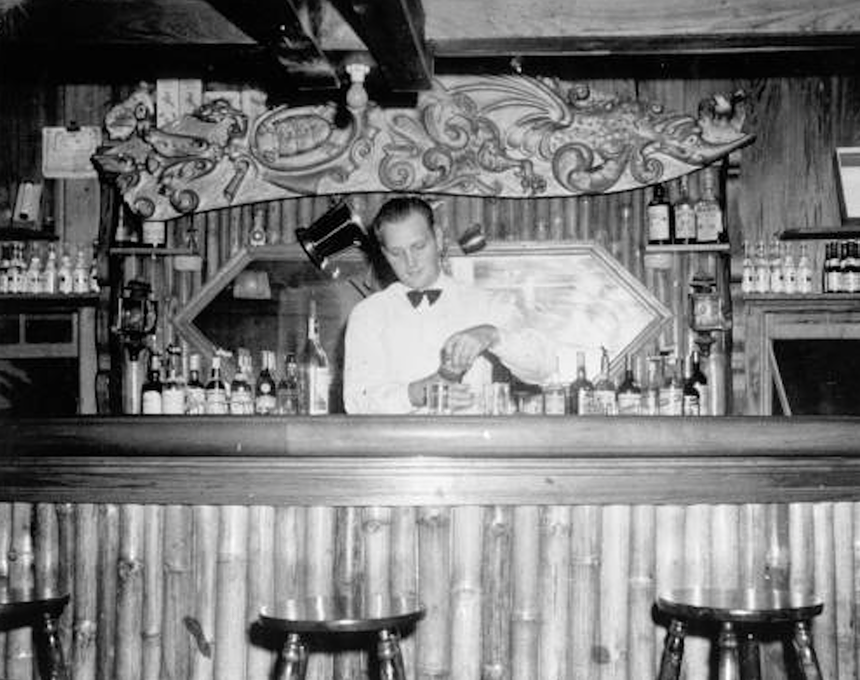 a black and white photograph of a man preparing drinks behind a bar