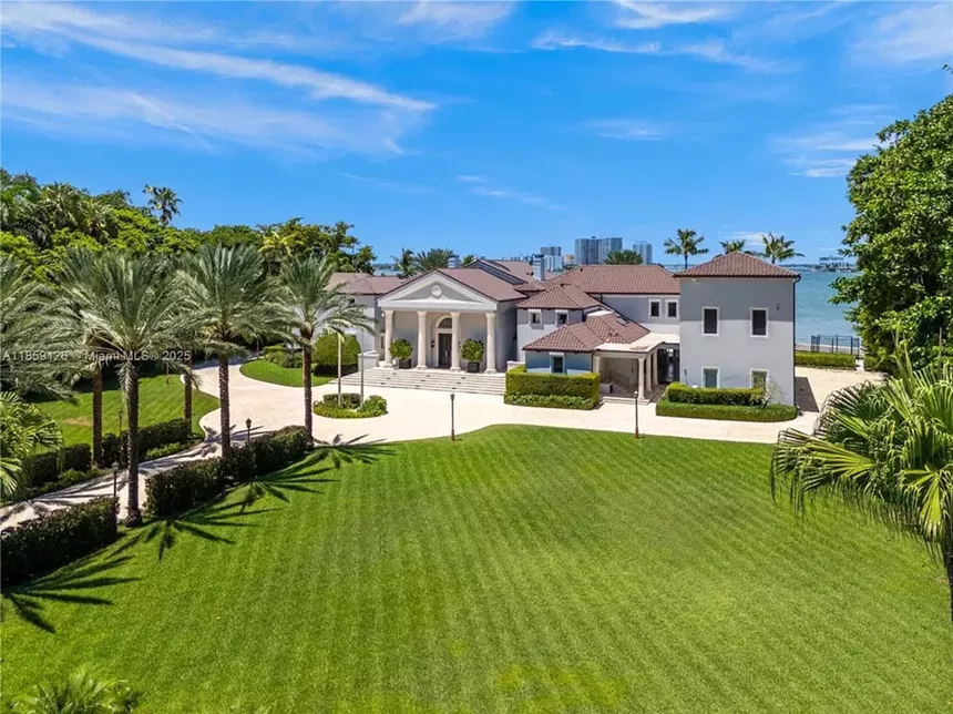 Pictured is a large front lawn with stripes cut into the grass and palm trees lining either side of a brick driveway, which leads up to an extravagant mansion.