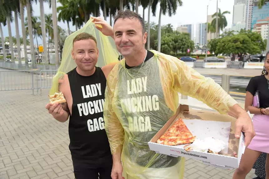 Two men wear rain ponchos and shirts reading, "Lady Fucking Gaga." The man on the right holds a pizza box with one remaining slice of pizza