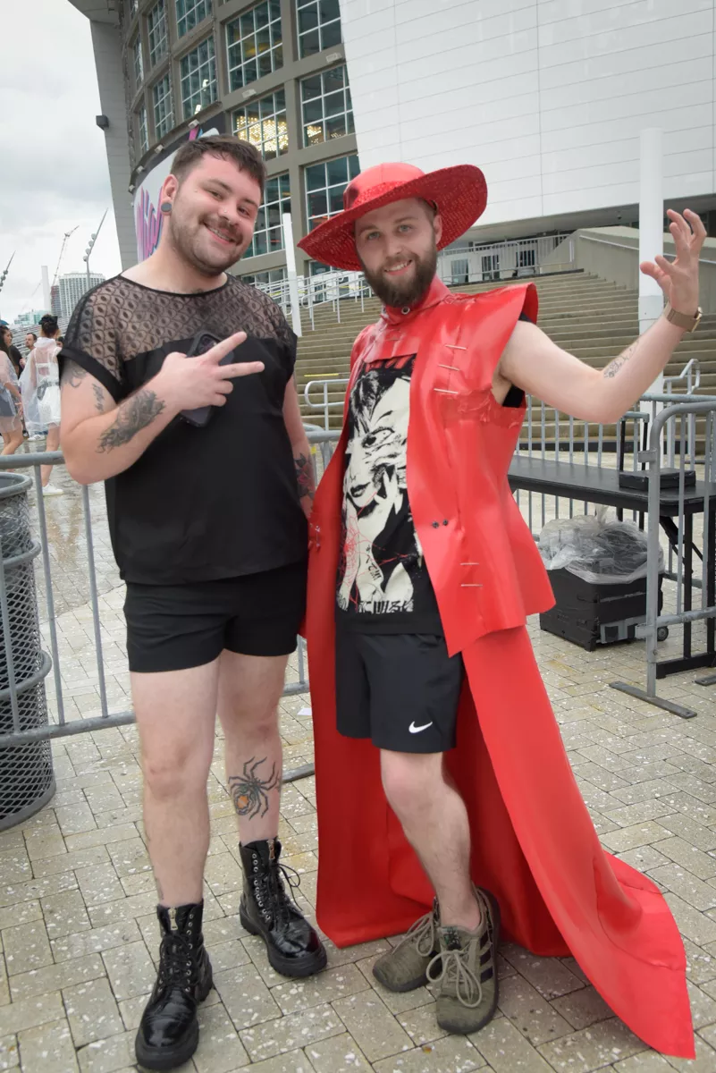 Two people stand in front of an arena. The one on the left wears a black blouse and shorts. The one on the right wears a graphic tee, a red fedora, and a plastic cape