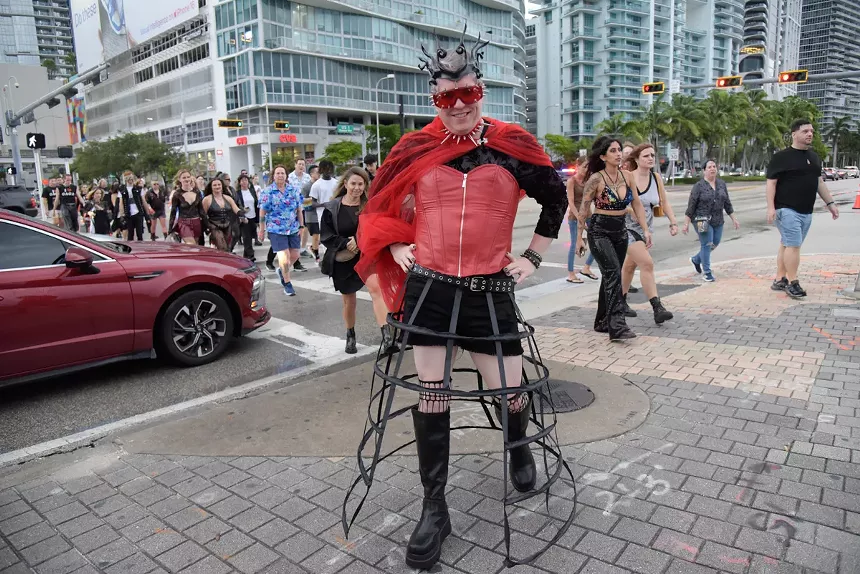 A person wears a red corset, studded choker, and cape with black shorts, and a frame for a sculpture skirt. They stand in front of a crosswalk with fans headed to a concert.
