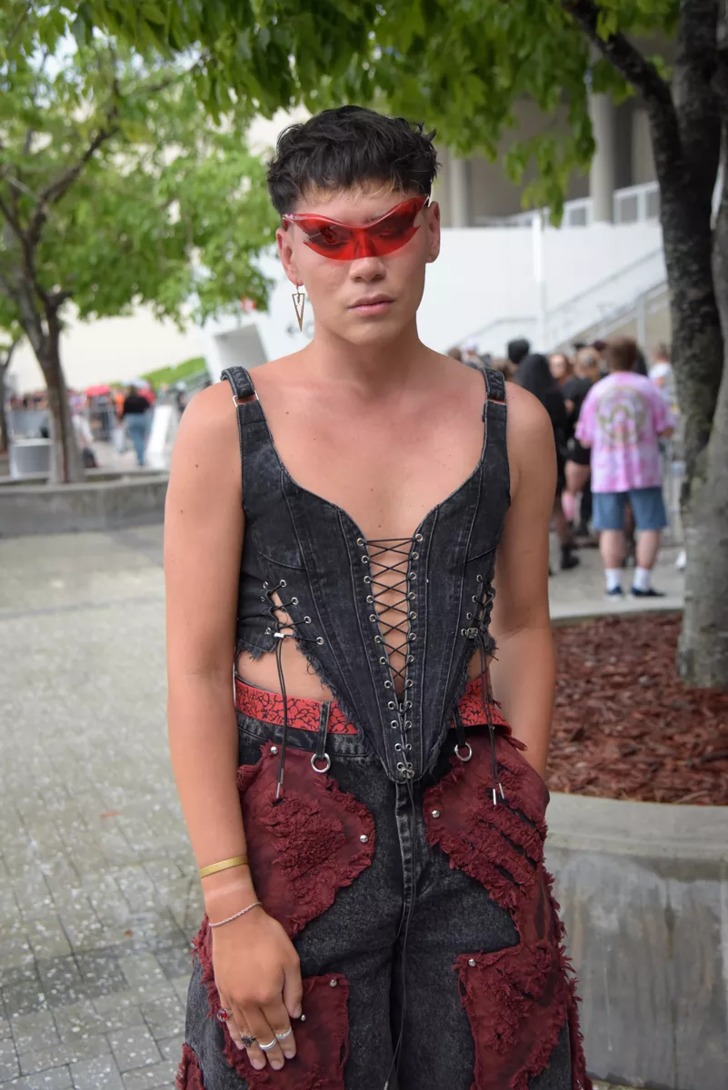 a person wears red graphic shades, a denim corset, and denim pants with maroon fabric overlay