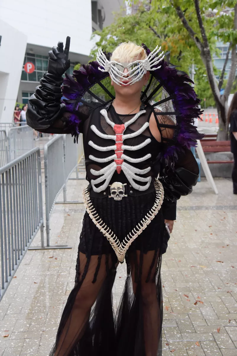A person wears shades made out of plastic skeleton hands, an elevated collar made of purple feathers, a black outfit adorned with a bedazzled sternum, and a skirt adorned with fake bones