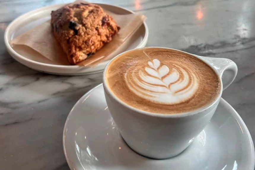 a latte on a glass saucer plate beside a fruity scone